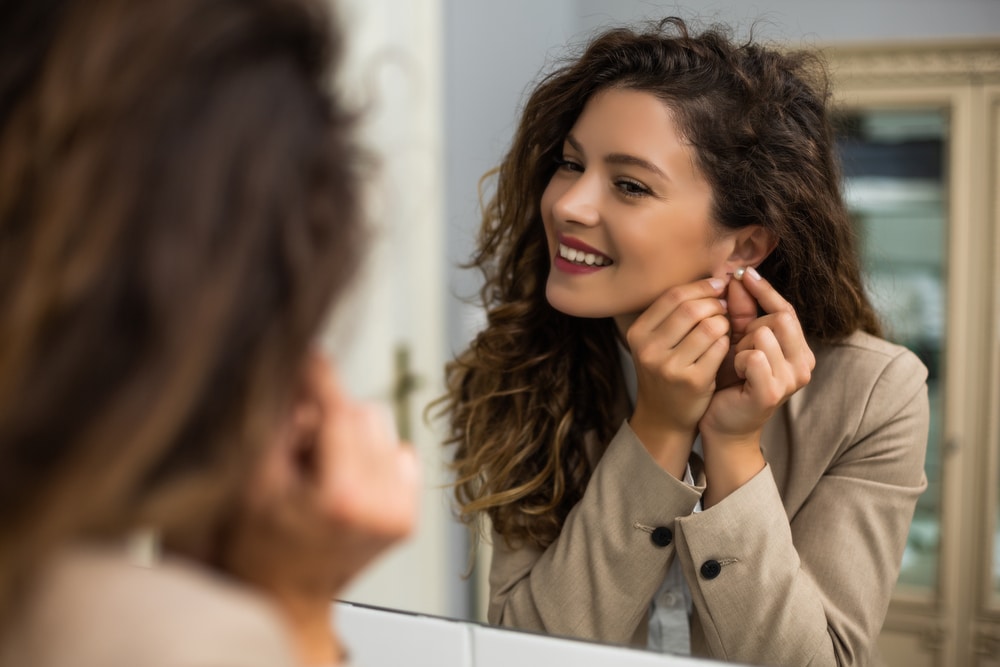 woman is putting earrings while looking in a mirror